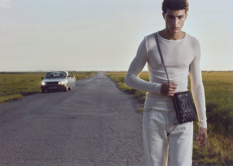 A young adult male model walks down a road holding a small black bag, with a car in the background, set against a rural landscape.