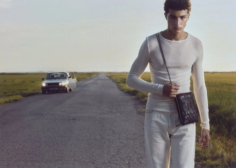 A young adult male model walks down a road holding a small black bag, with a car in the background, set against a rural landscape.