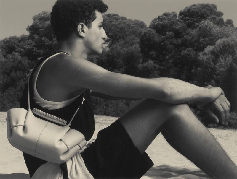Monochrome portrait of a male model with a shoulder bag sitting on a sandy beach, with a leafy background and natural lighting.
