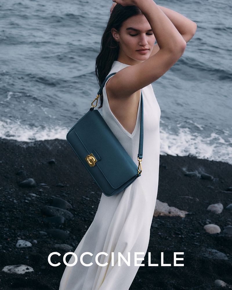Brunette fashion model in white dress holds a teal leather Coccinelle handbag on a rocky beach near ocean water with crashing waves.