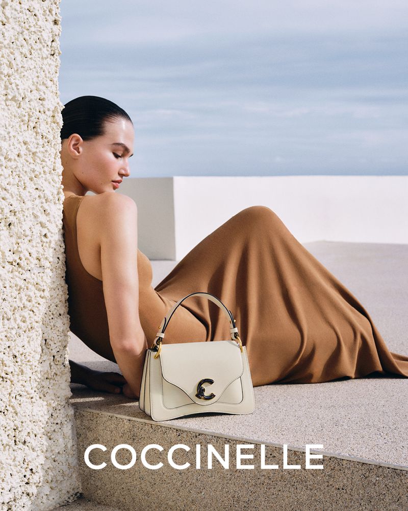 A tan-skinned woman in a camel dress poses near a cream-colored leather Coccinelle handbag against a blue sky background.