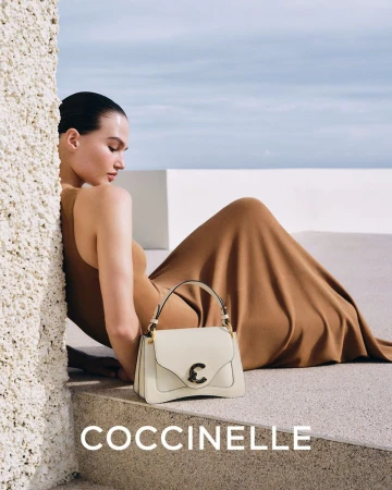 A tan-skinned woman in a camel dress poses near a cream-colored leather Coccinelle handbag against a blue sky background.