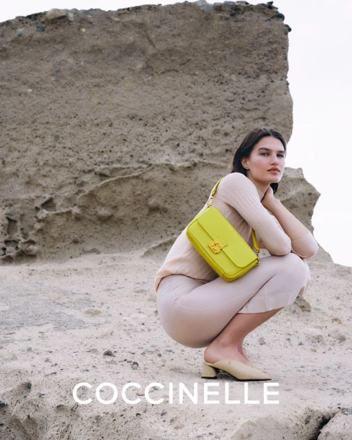 A woman wearing a light sweater and skirt is sitting on a rock and holding a yellow Coccinelle purse with a rocky background.