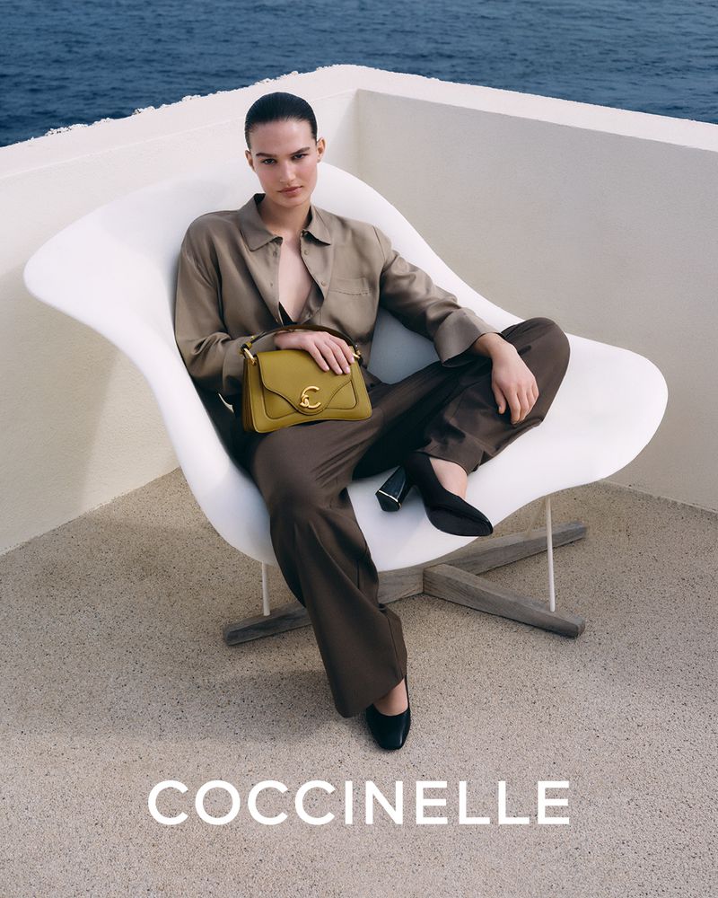 Model seated in a white sculptural chair holding an olive green handbag with ocean visible in the background