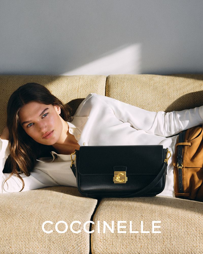 Brunette model lays on a couch with a Coccinelle black leather handbag. Indoor studio shot with neutral background and soft lighting.
