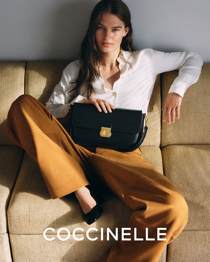 A woman poses on a couch holding a black leather purse. She wears a white blouse, brown pants, and black shoes in a fashion shot.