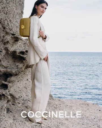 Woman in a cream-colored suit poses next to a rock formation, carrying a gold-colored Coccinelle handbag, with the ocean in the background.