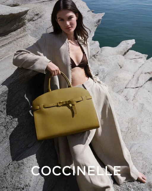 Brunette woman poses outdoors with olive-green Coccinelle handbag, wearing linen pants and jacket, on rocky terrain near ocean.