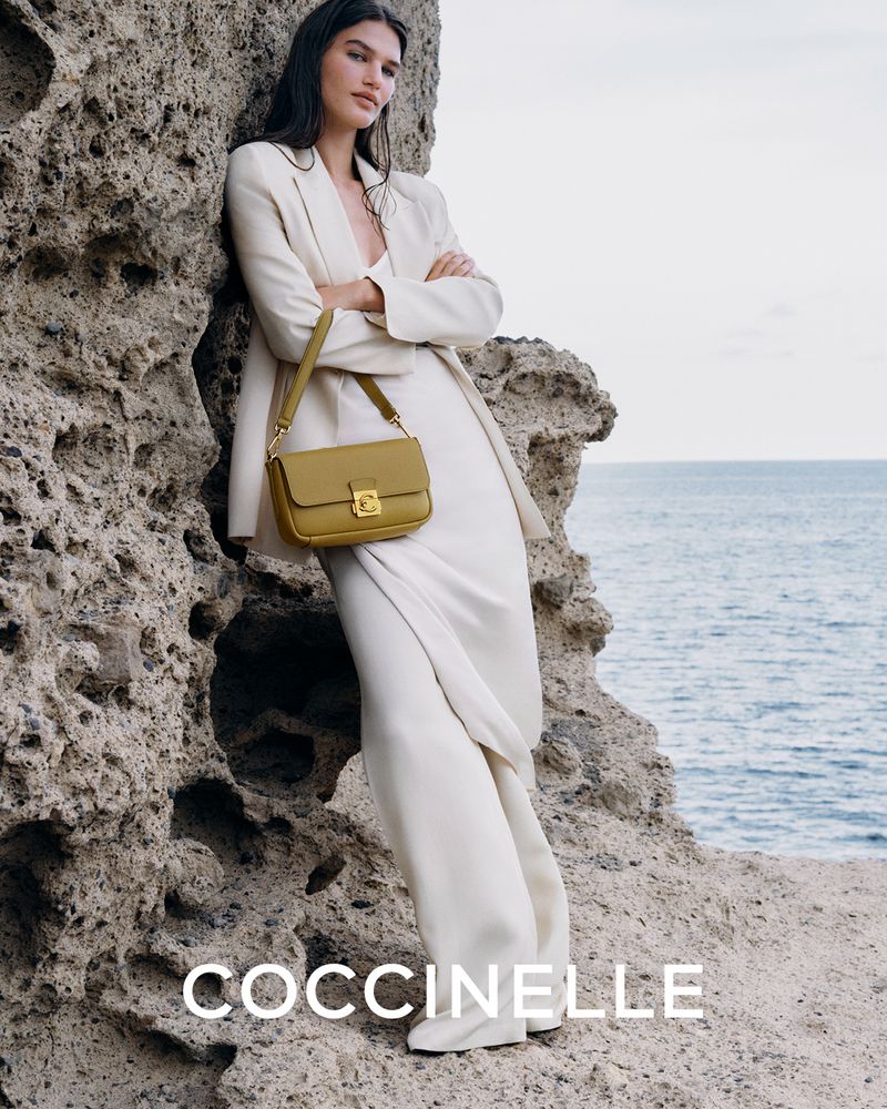 Woman in a beige suit and white shoes poses with a light-olive-colored Coccinelle handbag against a rocky coastal background.