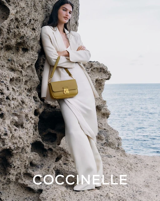 Woman in a beige suit and white shoes poses with a light-olive-colored Coccinelle handbag against a rocky coastal background.