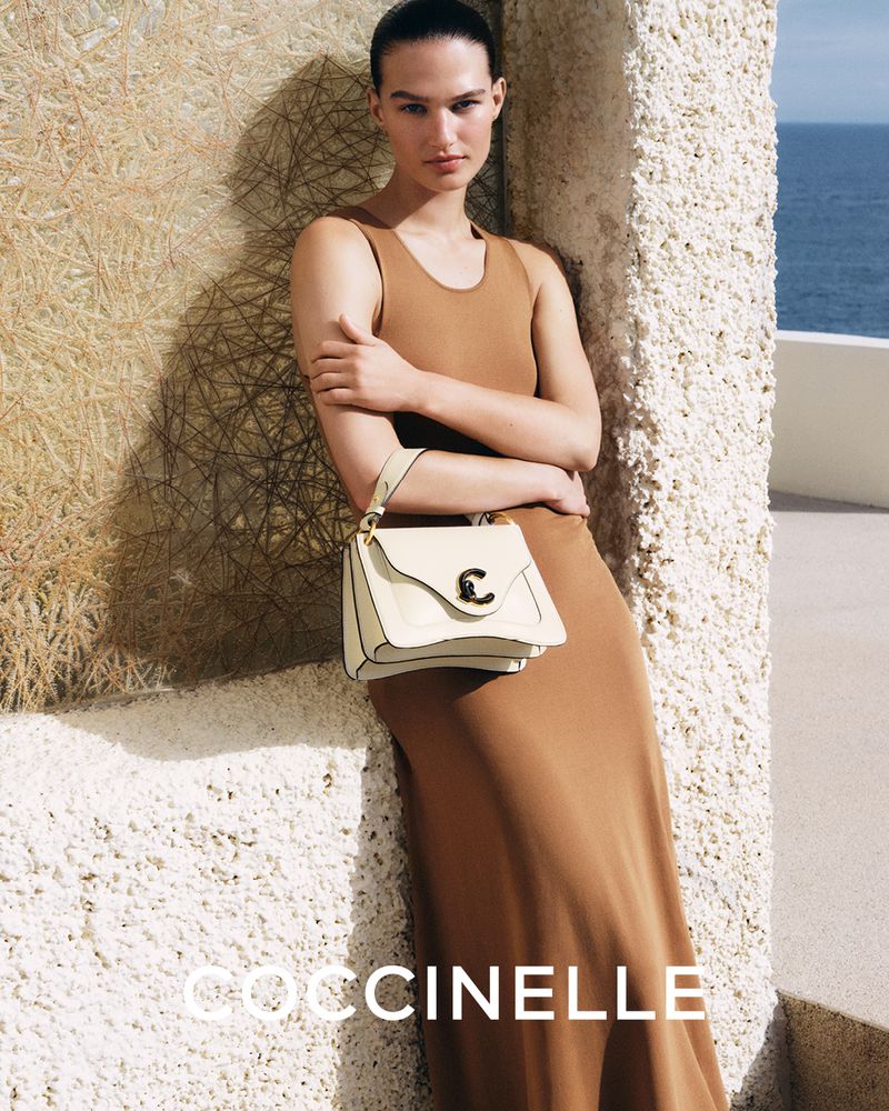 Woman in a brown dress poses with a cream-colored Coccinelle handbag against a textured wall with a view of the ocean in the background.