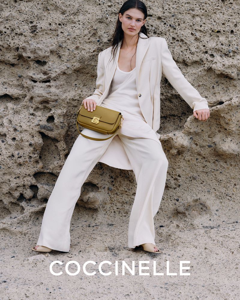 Fashion shot of a woman in a cream suit holding a green purse, posing against a textured rock background with the Coccinelle logo.