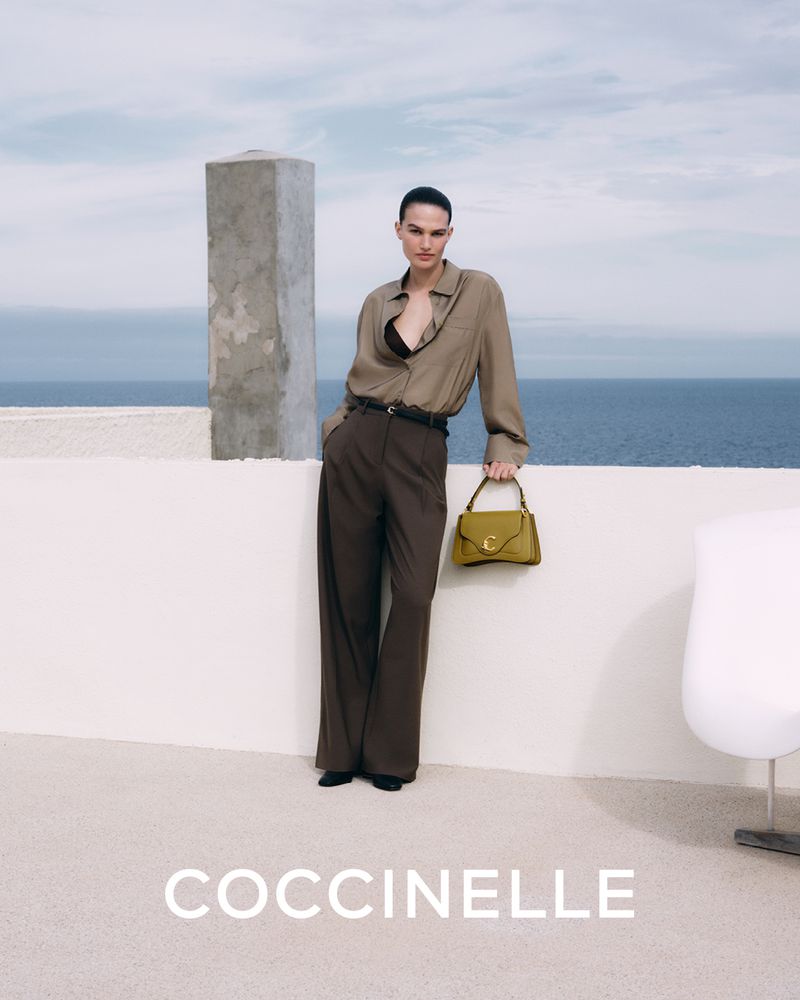 Fashionable woman in earth-tone outfit holding a purse outdoors with ocean backdrop. Coccinelle brand is visible in the image.
