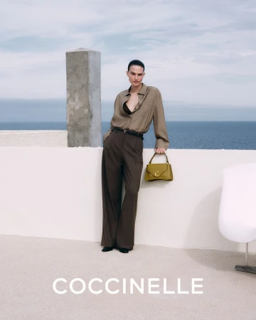 Fashionable woman in earth-tone outfit holding a purse outdoors with ocean backdrop. Coccinelle brand is visible in the image.