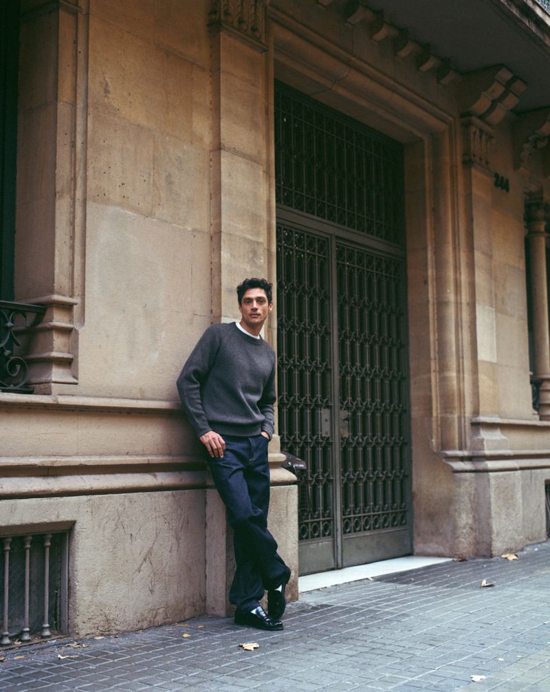 Man wearing a dark grey crewneck sweater leaning against a classical stone building exterior with tall double doors