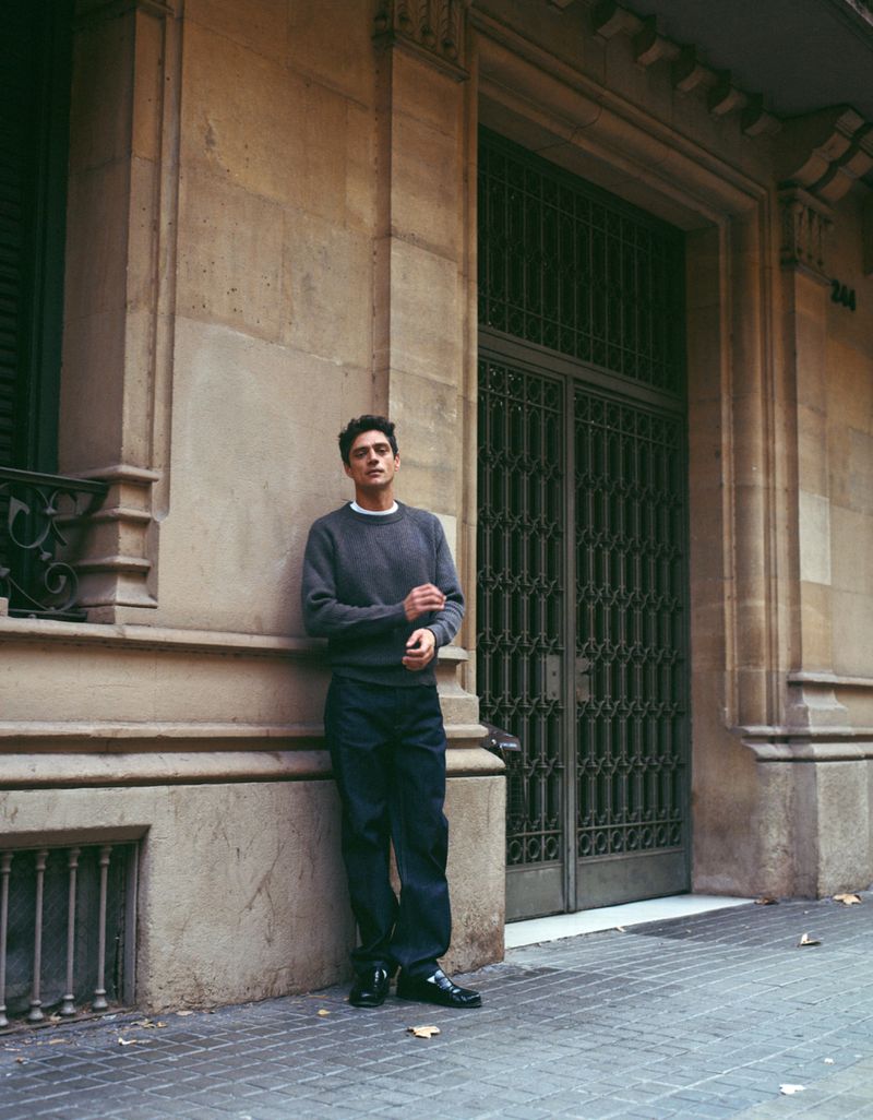 A man stands next to a stone building with a doorway; he is wearing casual clothing on a city street in an urban environment.