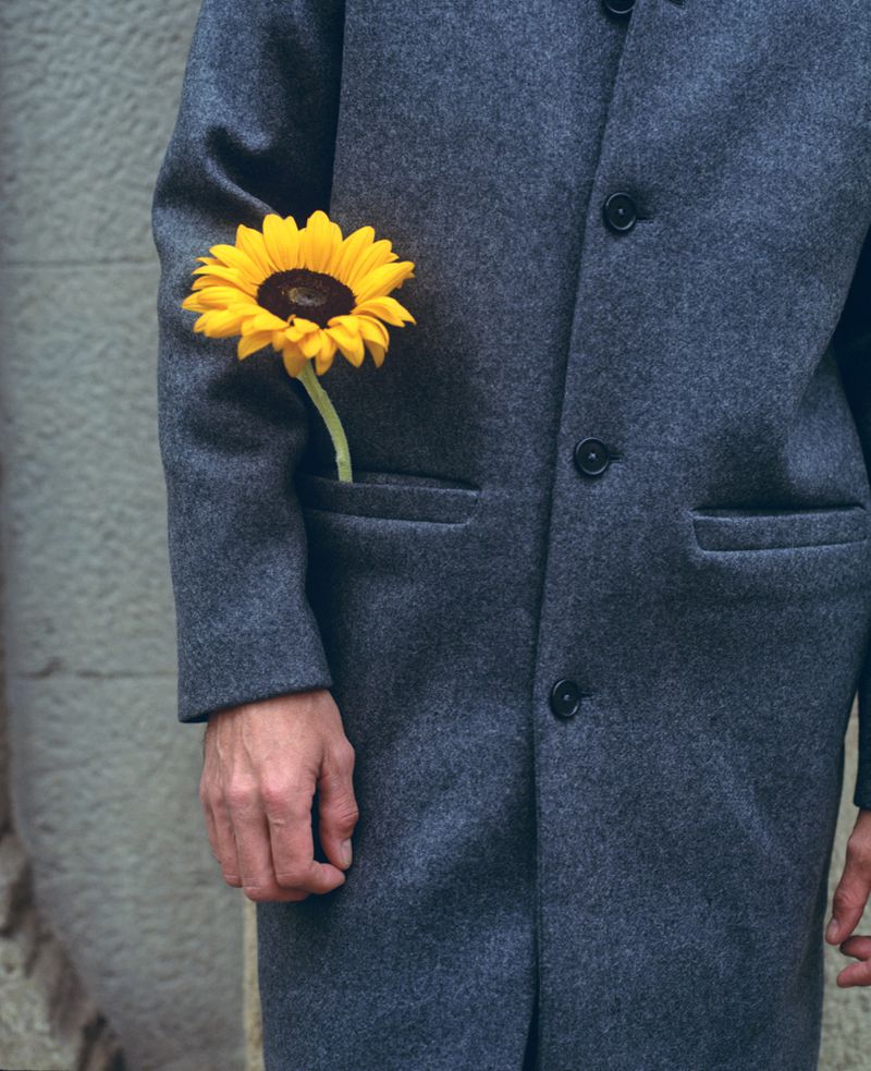 Close-up of an adult wearing a gray coat with a bright yellow sunflower in the pocket standing against a textured gray wall.