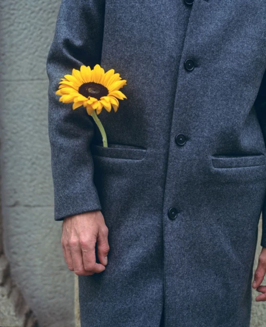 Close-up of an adult wearing a gray coat with a bright yellow sunflower in the pocket standing against a textured gray wall.
