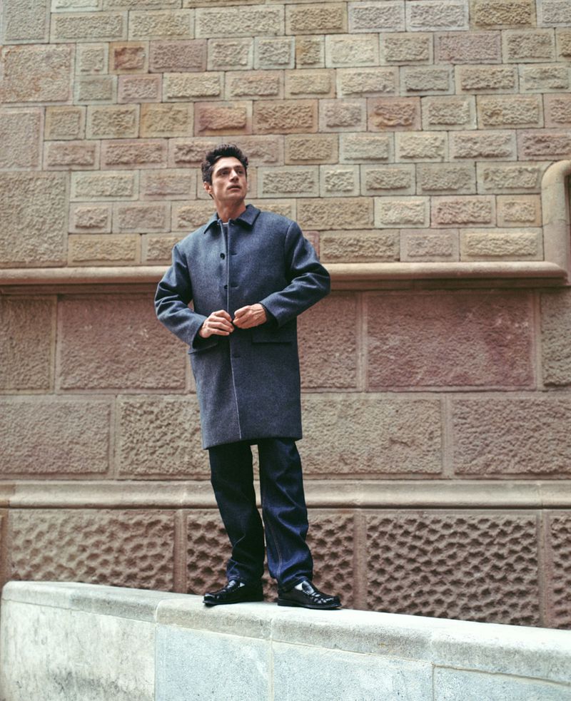 Adult man in a gray coat stands on a marble ledge in front of a tan brick wall, looking up and away, in an outdoor urban setting.