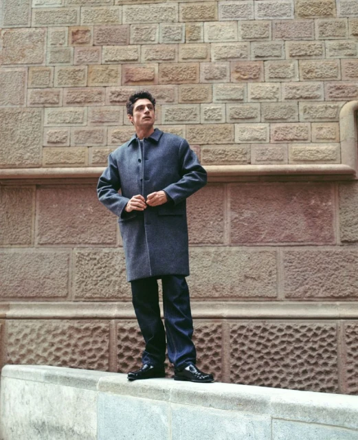 Adult man in a gray coat stands on a marble ledge in front of a tan brick wall, looking up and away, in an outdoor urban setting.