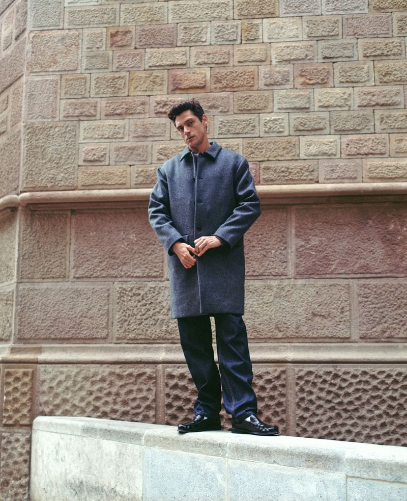 An adult male stands against a textured stone wall wearing a blue coat and jeans, with glossy black shoes, in an urban setting.
