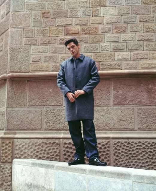 An adult male stands against a textured stone wall wearing a blue coat and jeans, with glossy black shoes, in an urban setting.