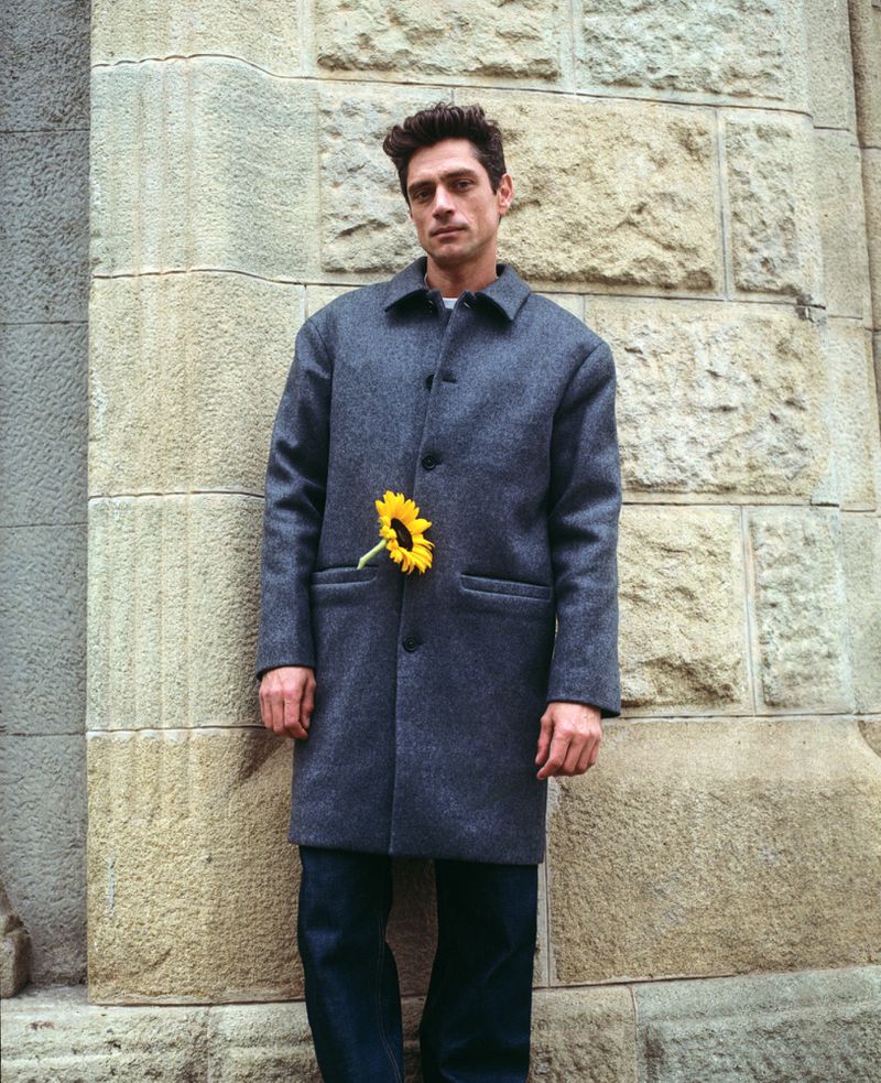 Man in grey coat stands against stone wall with a sunflower in his pocket, portrait shot, cool color palette, minimalist fashion.
