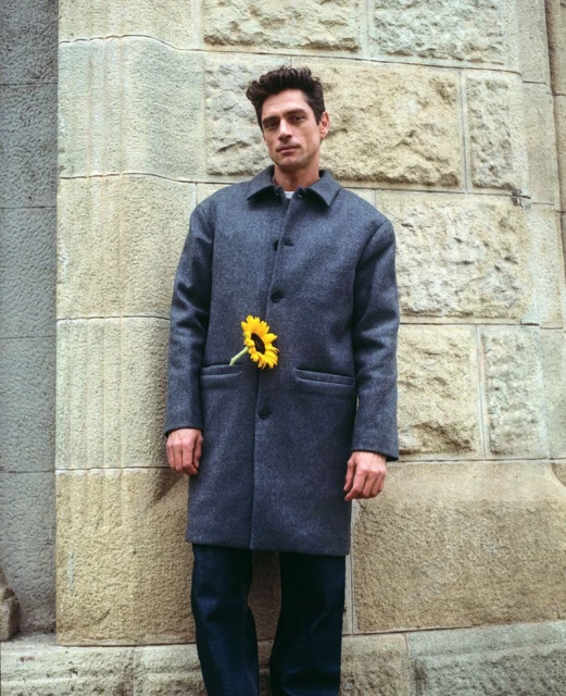 Man in grey coat stands against stone wall with a sunflower in his pocket, portrait shot, cool color palette, minimalist fashion.