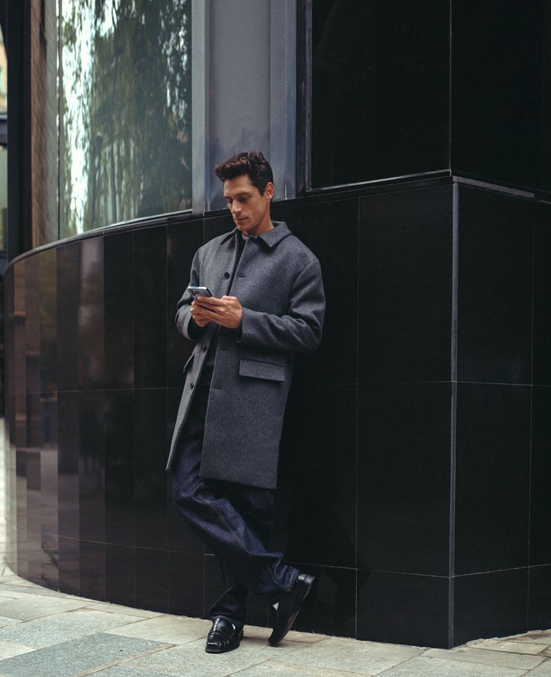 Adult male in a gray coat and dark jeans leaning against a modern, black tiled building while using a smartphone in an urban environment.