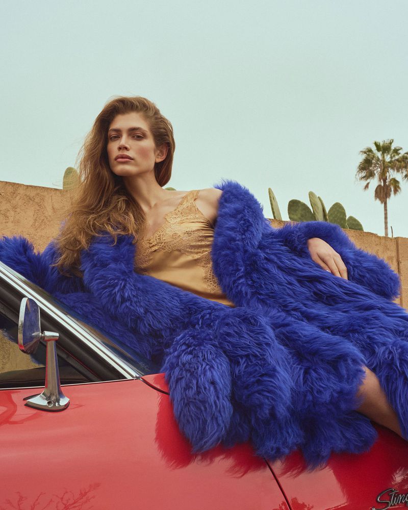 Adult female fashion model in gold dress and blue fur coat laying on a red vintage car under bright, overcast sky in outdoor setting.