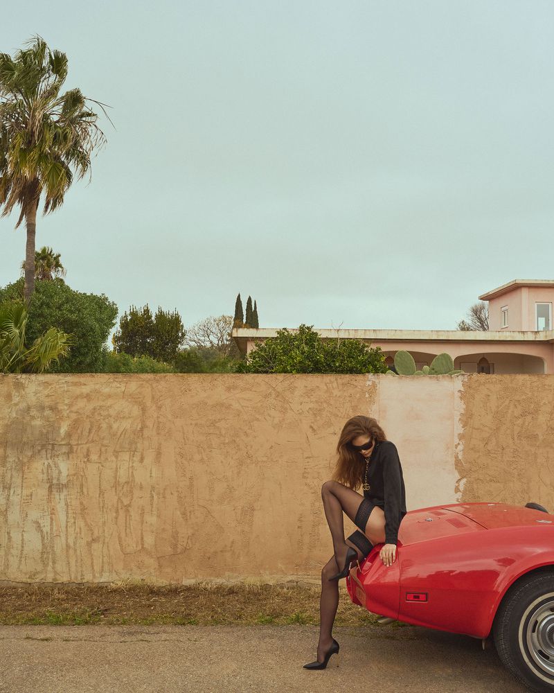 Woman in hosiery poses on a red sports car near a tan wall, under a cloudy sky with palm trees in the background, outdoor setting.
