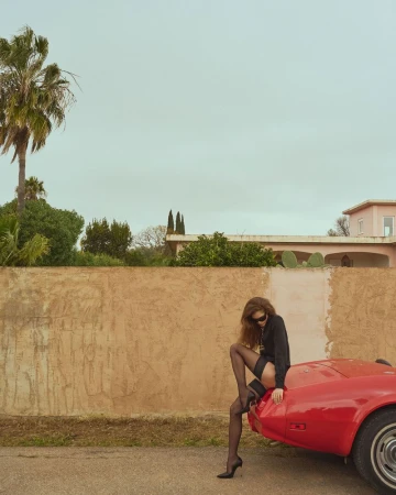 Woman in hosiery poses on a red sports car near a tan wall, under a cloudy sky with palm trees in the background, outdoor setting.