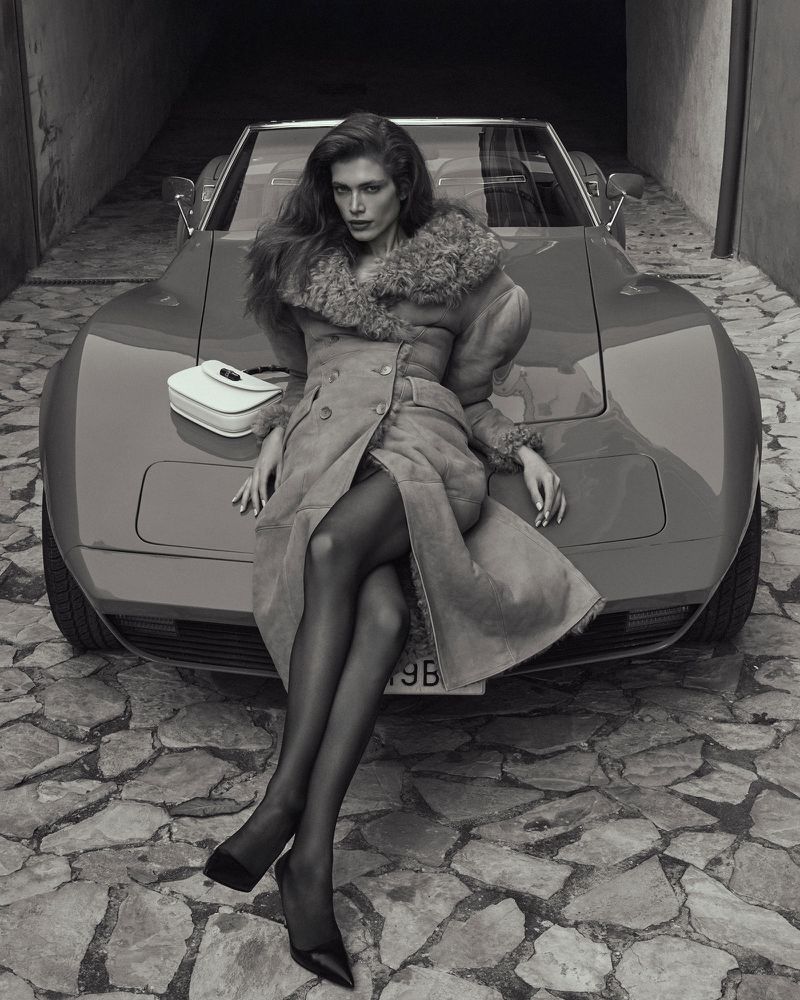 A stylish woman in a coat poses on a red Corvette with a purse. She is wearing black tights and heels in an urban setting.