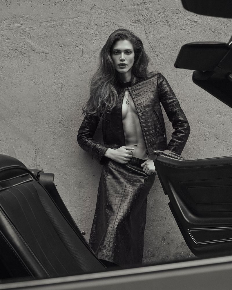 Black-and-white fashion shot of a woman in a leather jacket and skirt, standing next to a car, leaning against a textured wall.