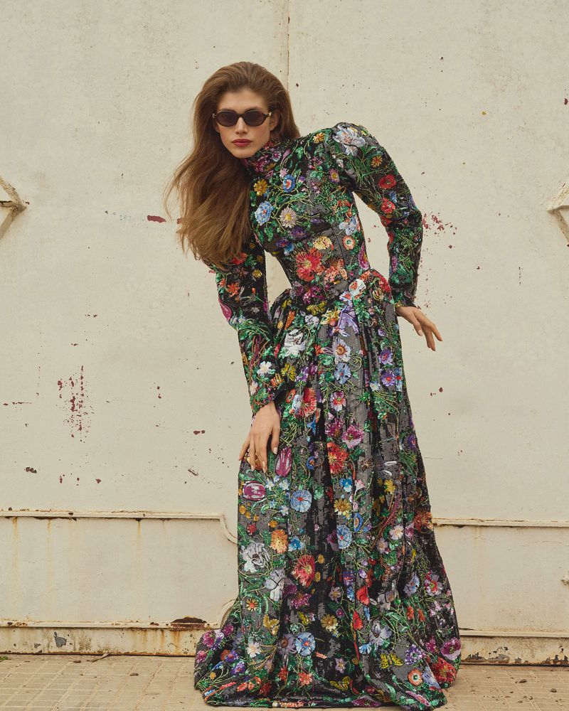 A single adult female model wears a long-sleeved floral gown and sunglasses, posing against a weathered wall in an outdoor setting.
