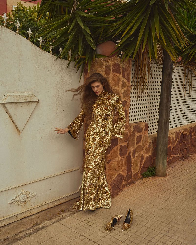 Model wearing a glittering gold dress posing near a white gate and stone wall outdoors under palm fronds
