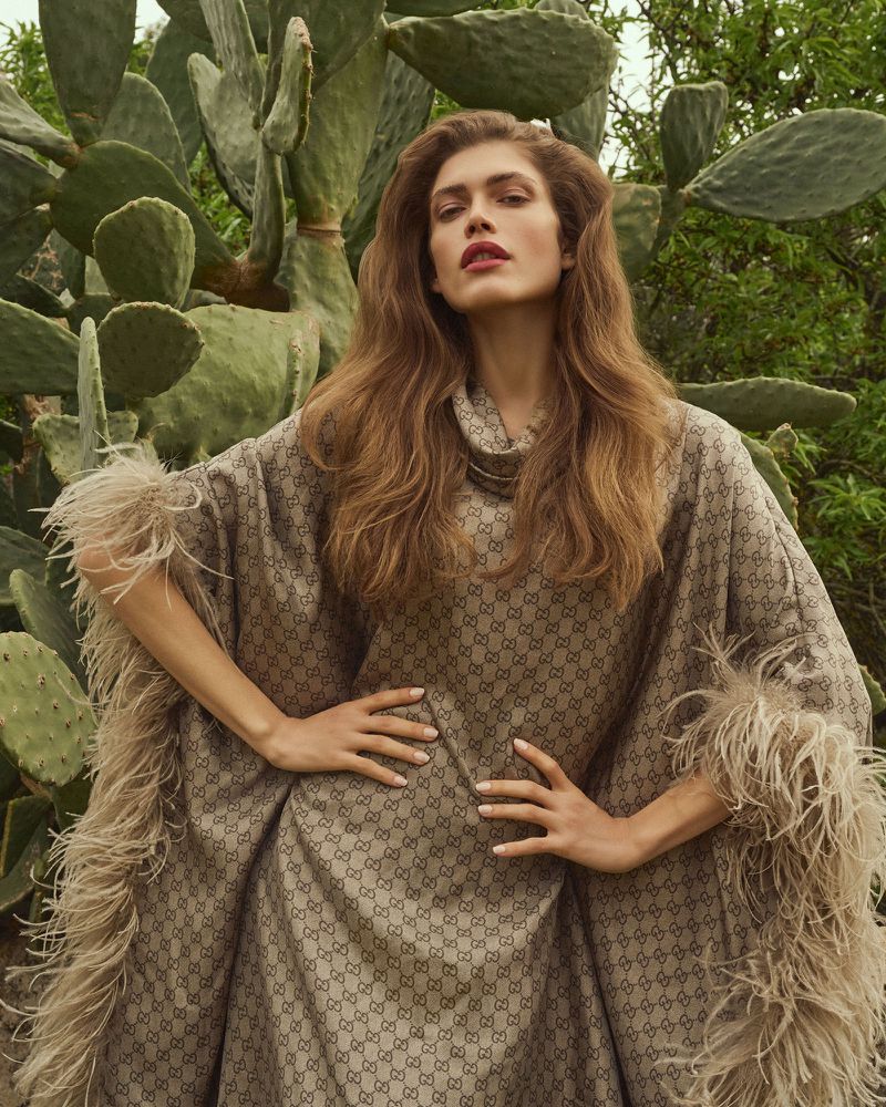 Fashion shot of a woman with long brown hair wearing a patterned poncho with feathered edges, standing outdoors in front of cacti.