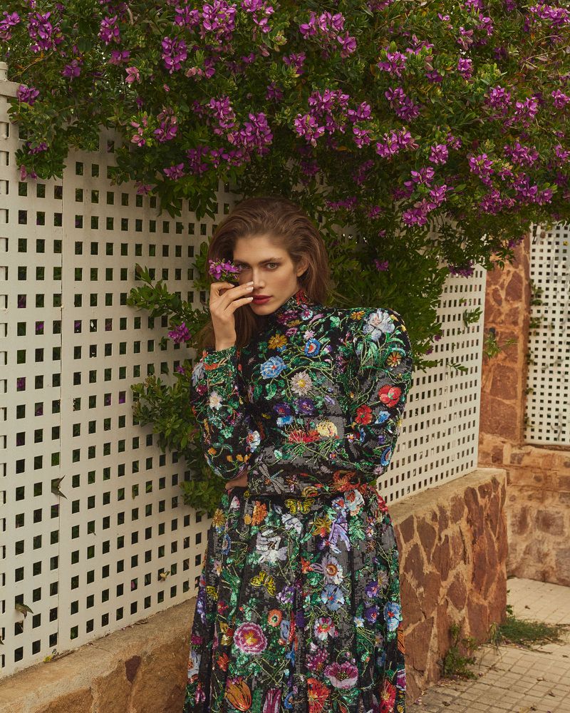 A woman poses in a long floral dress against a white lattice fence with purple flowers, capturing a blend of fashion and nature.