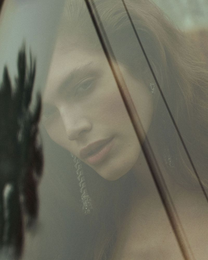 Close-up portrait of a woman with brown hair and dangling earrings, reflected and obscured by glass, with a tree silhouette.
