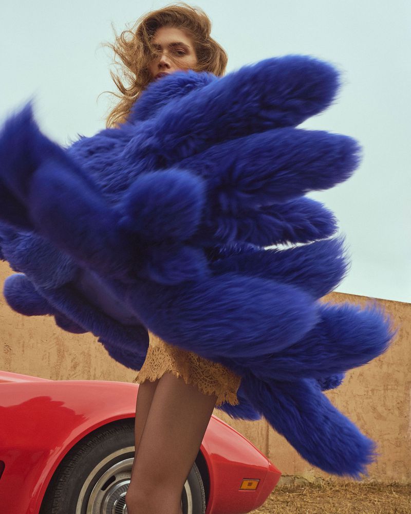 Woman in a cobalt fur coat with a red sports car. She poses outdoors, against a beige wall and light blue sky in the background.