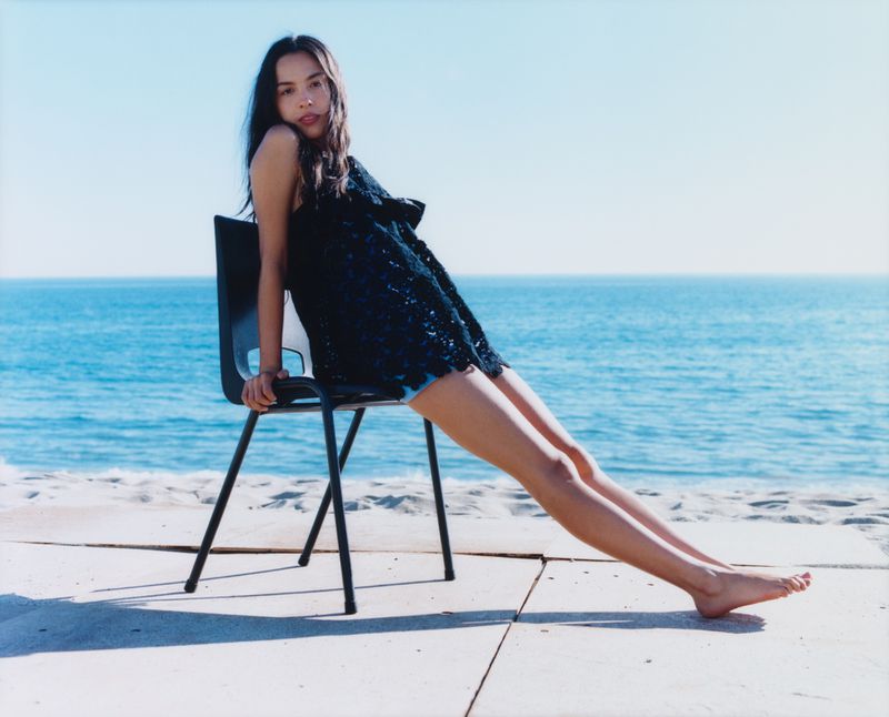 Young woman in dark dress poses on black chair near the ocean on a sunny day. Ocean backdrop, neutral colored platform. 