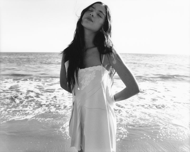 Woman stands in a white dress with her hands behind her back on a beach with waves in a black and white outdoor fashion shot.