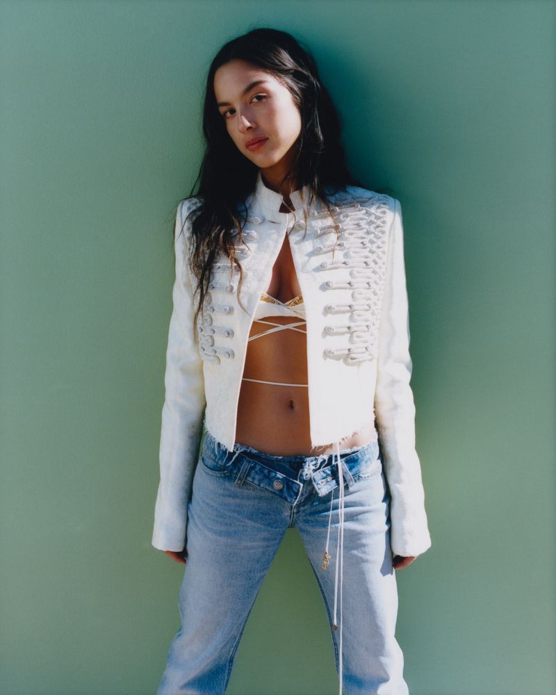 A woman with long dark hair poses against a green wall wearing a white jacket, light blue jeans, and a beige lace top with strings.