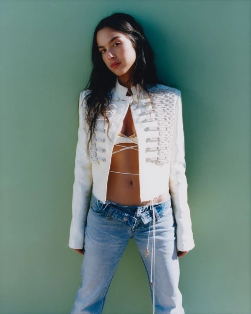 A woman with long dark hair poses against a green wall wearing a white jacket, light blue jeans, and a beige lace top with strings.