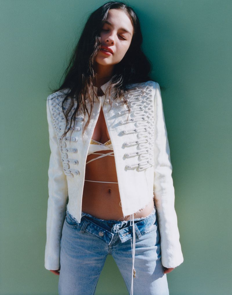 A woman poses against a green wall, wearing a white jacket and low-rise jeans, in a fashion shot with a bright outdoor lighting.