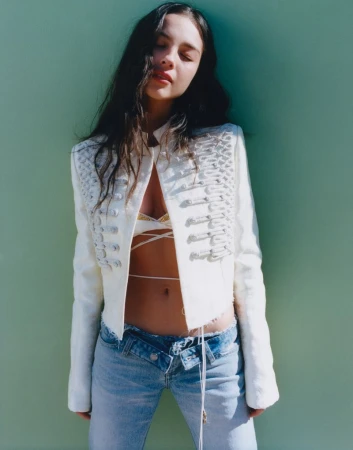 A woman poses against a green wall, wearing a white jacket and low-rise jeans, in a fashion shot with a bright outdoor lighting.