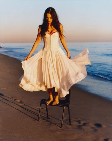 Woman in flowing white dress stands barefoot on a chair at the beach at sunrise. Ocean waves and sandy shore are visible.