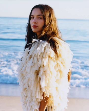 Portrait of a young adult female with long brown hair wearing a fringed jacket, posing on a beach with blue ocean waves in the background.