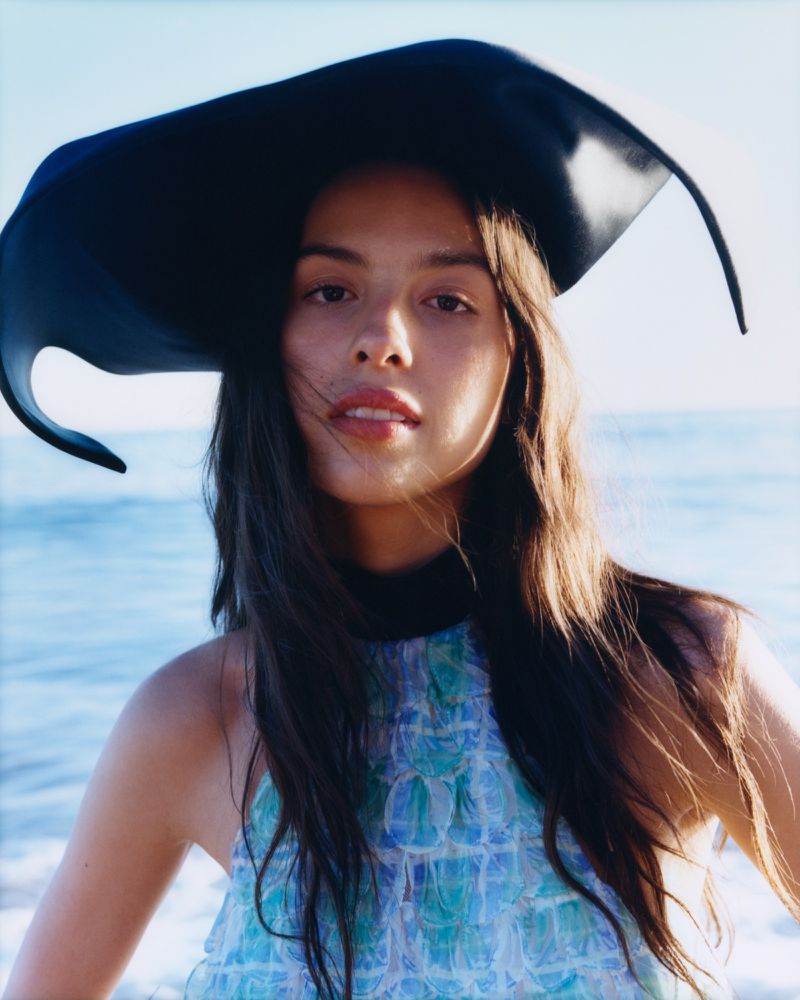 Woman in a blue and green halter top and wide-brimmed black hat poses outdoors with blurred ocean background in a portrait shot.