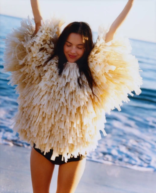 A woman in a fringed beige top poses on a beach with arms raised, featuring a blue ocean backdrop in an outdoor setting.